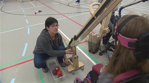 Josée Duquette se tient dos au jeu et manipule une rampe de lancement pour permettre à sa fille de lancer les boules. Josée Duquette se tient dos au jeu et manipule une rampe de lancement pour permettre à sa fille de lancer les boules.