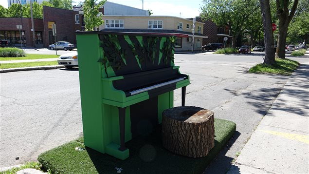 Photo : piano public de Limoilou. Vert forêt, une tranche d'arbre comme siège. Au bord d'une stationnement.