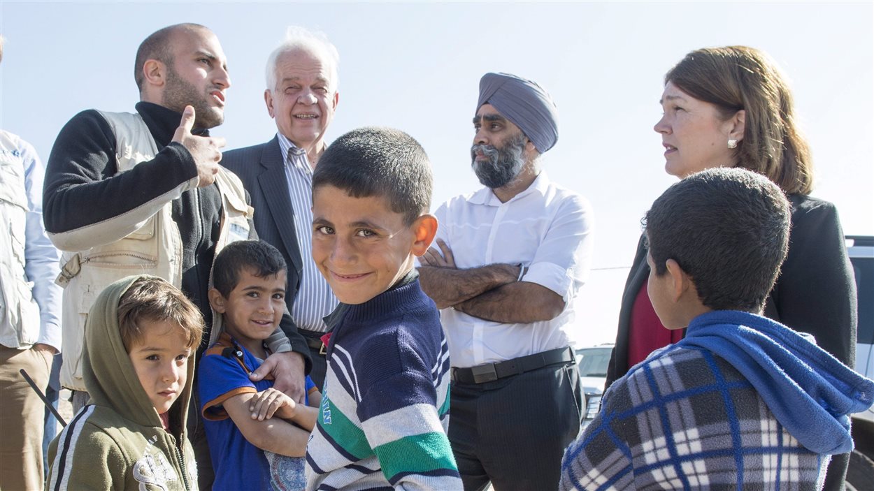 De jeunes Syriens entourent le ministre de l'Immigration, des Réfugiés et de la Citoyenneté John McCallum, le ministre de la Défense Harjit Sajjan et la ministre de la Santé Jane Philpott dans un camp de réfugiés en Jordanie le 29 novembre 2015. De jeunes Syriens entourent le ministre de l'Immigration, des Réfugiés et de la Citoyenneté John McCallum, le ministre de la Défense Harjit Sajjan et la ministre de la Santé Jane Philpott dans un camp de réfugiés en Jordanie le 29 novembre 2015.